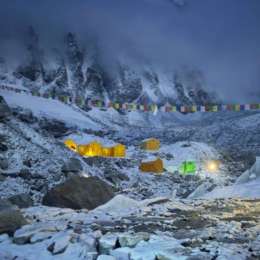Snow-covered mountain peaks at Everest Base Camp with small glowing yellow and green tents or cabins on a rocky slope, plus colorful prayer flags stretching across the scene.