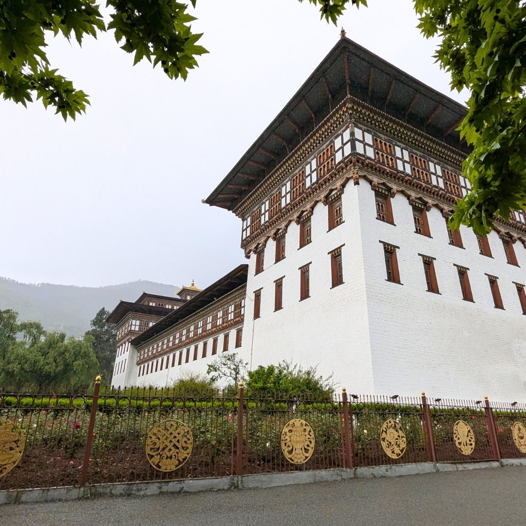 Traditional Bhutanese building with white walls and wooden accents, surrounded by greenery.